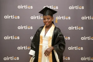 An Airtics graduate smiles in front of the Airtics photo backdrop